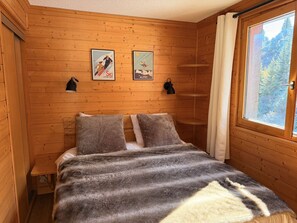 A wooden cabin room with a bed, two wall-mounted lamps, and a window with a view of trees.