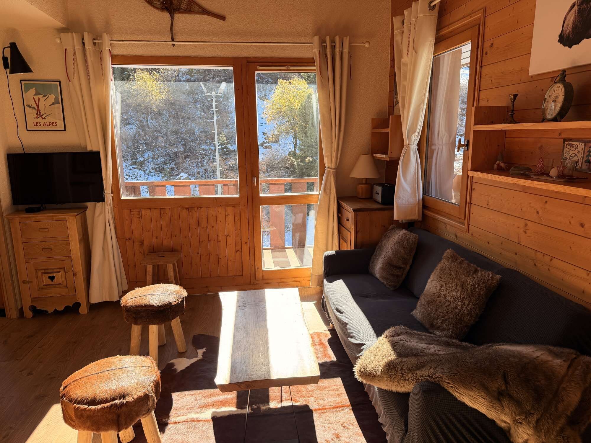 A cozy cabin interior with a wooden floor, a sofa with fur throw, a television, and a window with curtains.