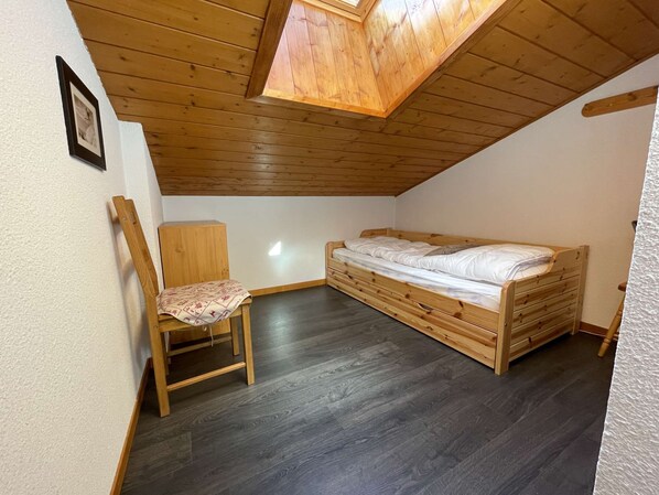 A small, single-room apartment with a sloped ceiling, a wooden bed, a chair, and a small wooden cabinet.