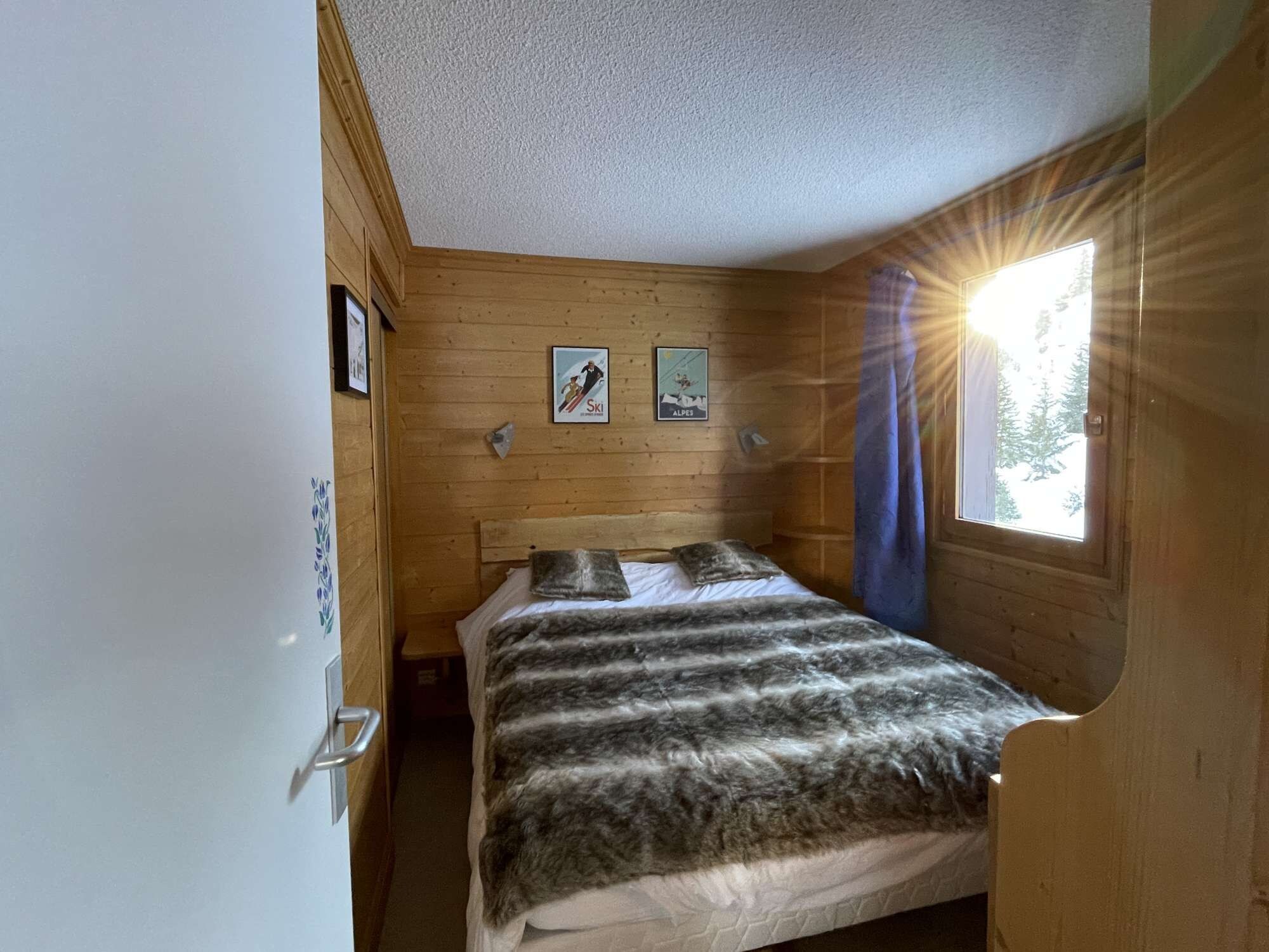 A small room with a bed, wooden walls, a window with a curtain, and two framed pictures on the wall.