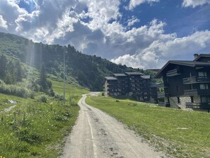 Un chemin de gravier mène à un ensemble d’immeubles modernes à plusieurs étages, dotés de balcons, et qui sont entourés d’une végétation luxuriante, avec une chaîne de montagnes en toile de fond.