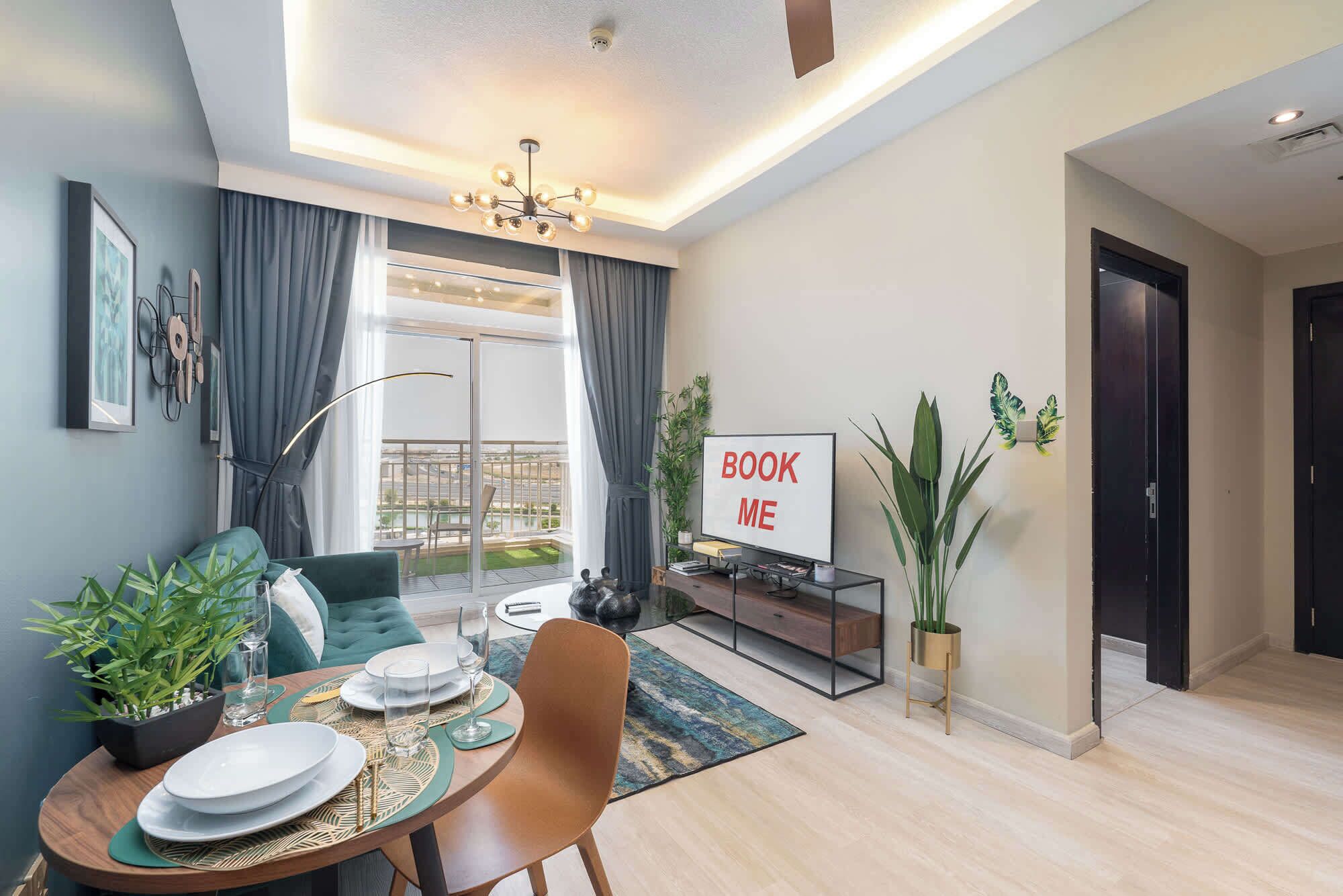 En moderne stue med spiseplads, et stort tv der viser "BOOK ME", og en balkon med udsigt.