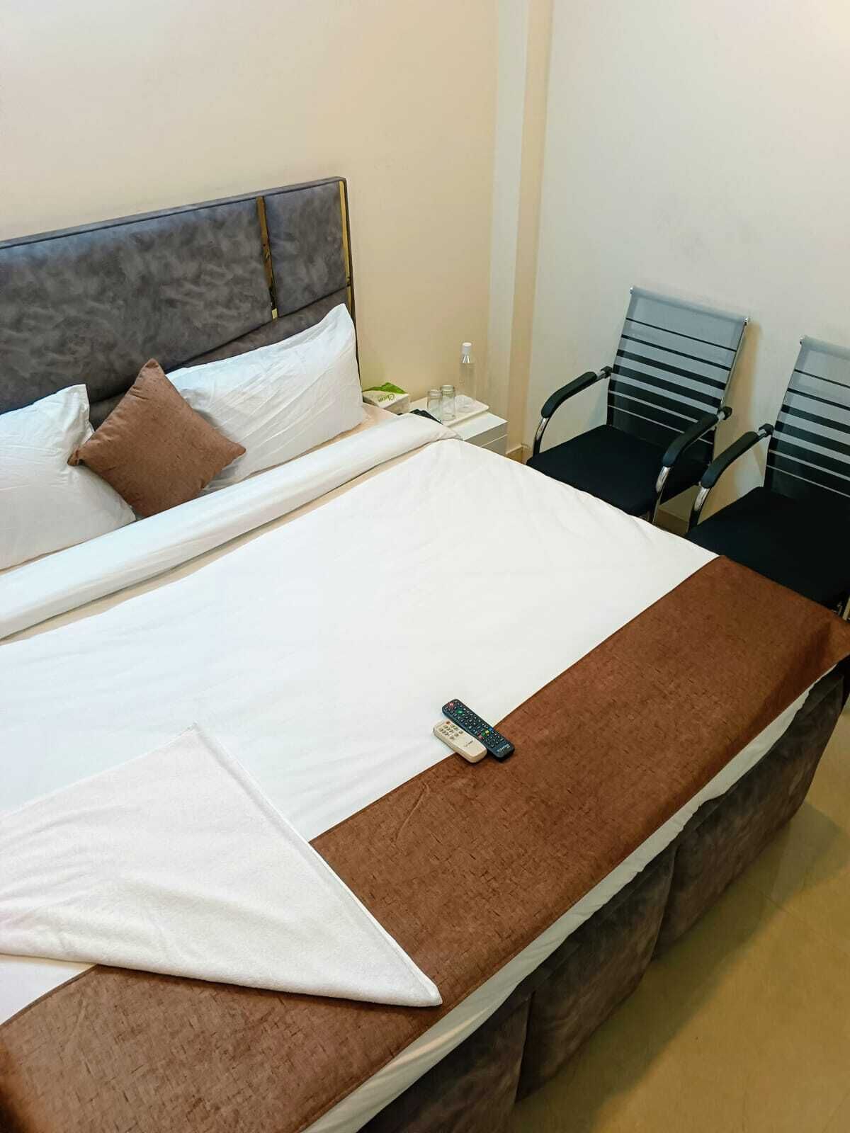 A neatly made bed with a brown throw blanket and pillows, a remote control on the bed, and two chairs against the wall.