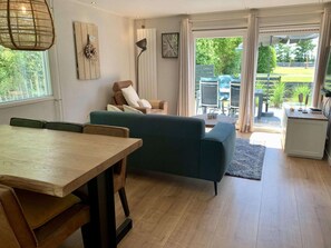 A modern living room with a wooden dining table, a green sofa, and a large window offering a view of the outdoors. - Bungalow for 4 Persons With South-facing Terrace (Sint Maartensvlotbrug)