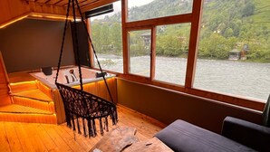 Een houten interieur met een groot raam met uitzicht op een rivier, een hangstoel en een bank.