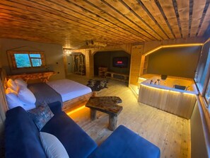 A cozy wooden interior with a bed, sofa, and a small table. A TV is mounted on the wall, and a fireplace is visible in the background.