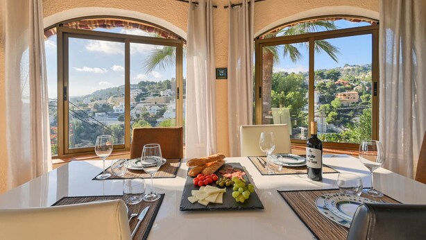 A dining table with a cheese board, wine, and bread, set in a room with large windows overlooking a scenic landscape. - Villa Las Brisas Benissa Costa (Benissa)
