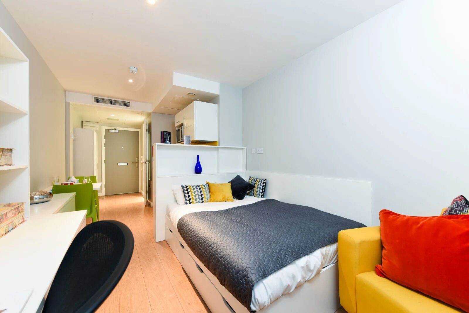A modern bedroom with a bed, a desk, and a chair. There is a kitchenette area in the background.