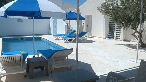 Une piscine entourée de chaises longues et de parasols, le tout bordé de murs blancs et d’un arbre.