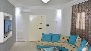Un salon avec un canapé, un téléviseur, un luminaire au plafond et une porte.