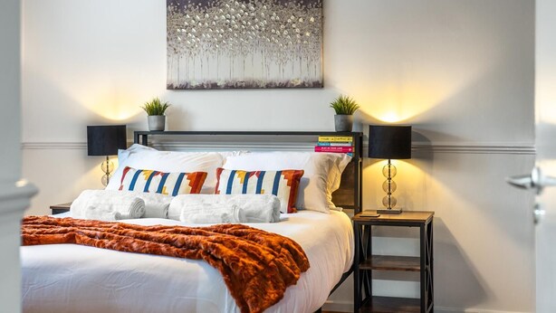 Une chambre d’hôtel moderne avec un lit soigneusement fait, une grande peinture abstraite au-dessus du lit, deux tables de chevet avec des lampes et une petite plante en pot sur chaque table.