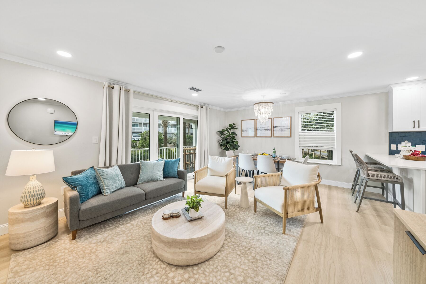 A modern living room with a grey sofa, round coffee table, and wicker chairs. There is a dining area with a table and chairs, and a kitchen area with a counter and cabinets.