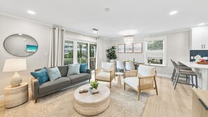 Uma sala de estar moderna com um sofá cinzento, mesa de centro redonda e cadeiras de vime. Há uma área de jantar com mesa e cadeiras, e uma cozinha com bancada e armários.