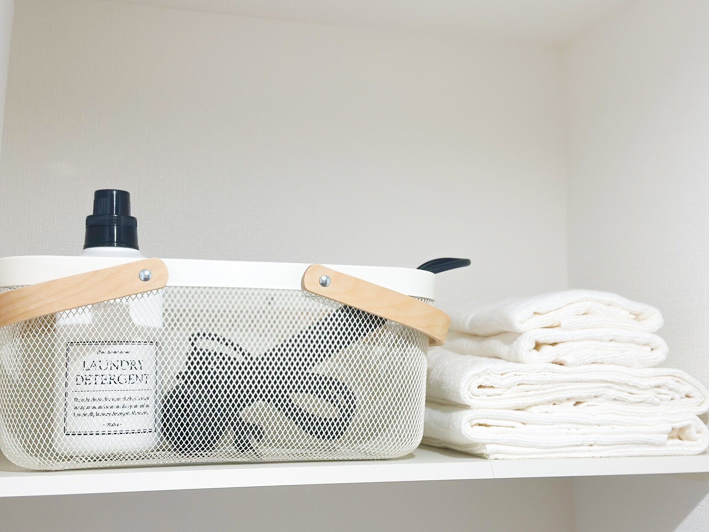A laundry basket with a mesh front and wooden handles, placed on a shelf next to folded white towels.