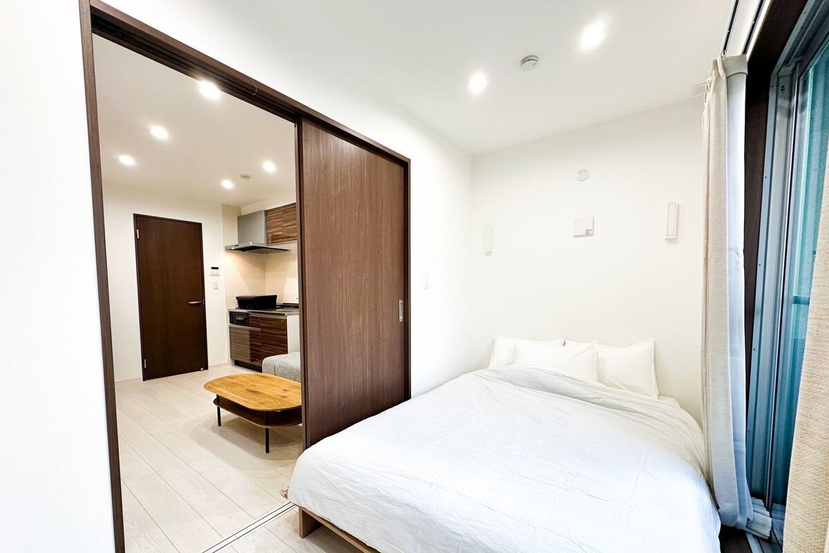A modern bedroom with a large bed, a wooden bench, and a kitchenette in the background.