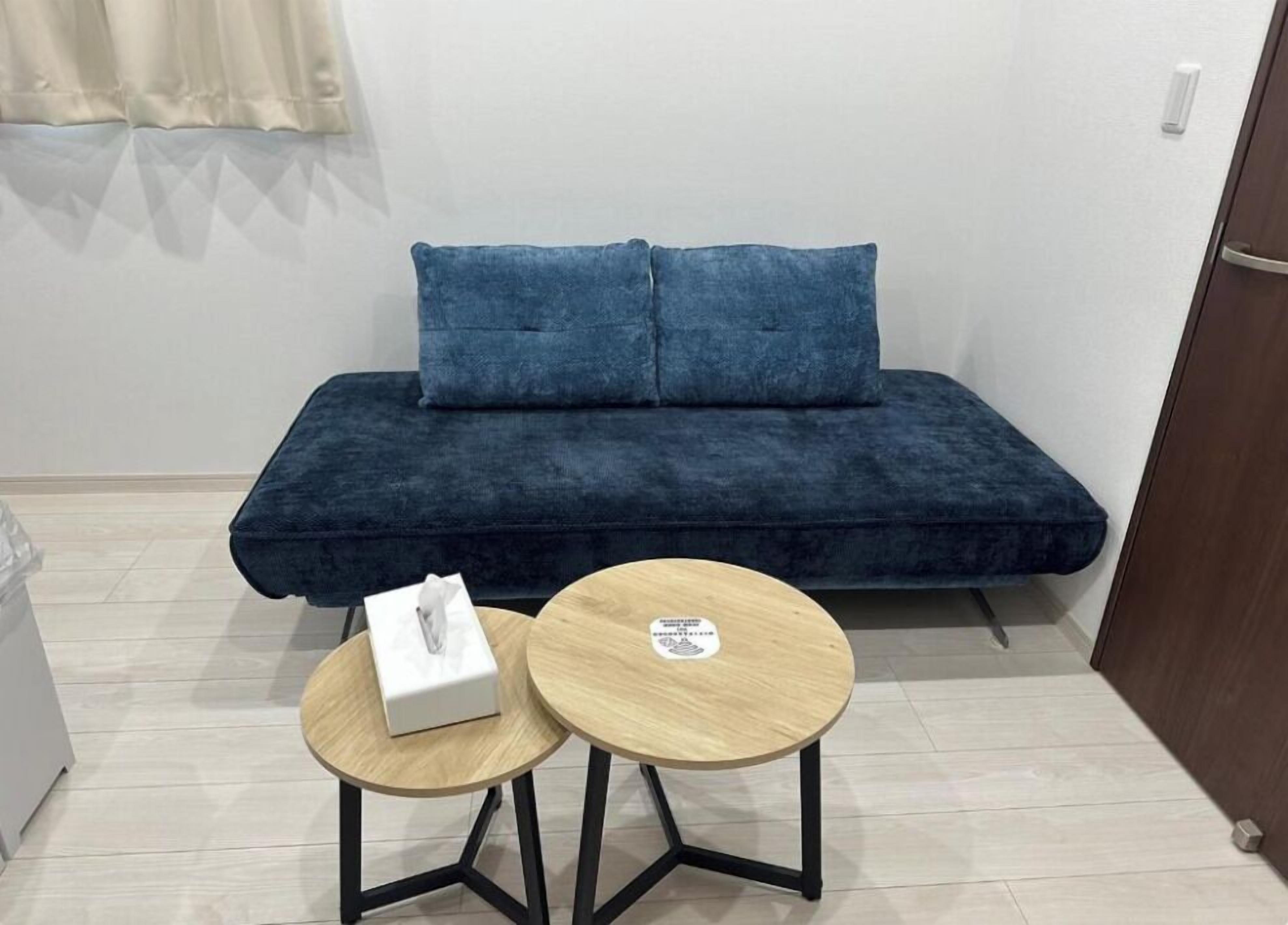 Una habitación pequeña con un sofá azul oscuro, dos mesas de madera y una caja blanca sobre una de ellas.
