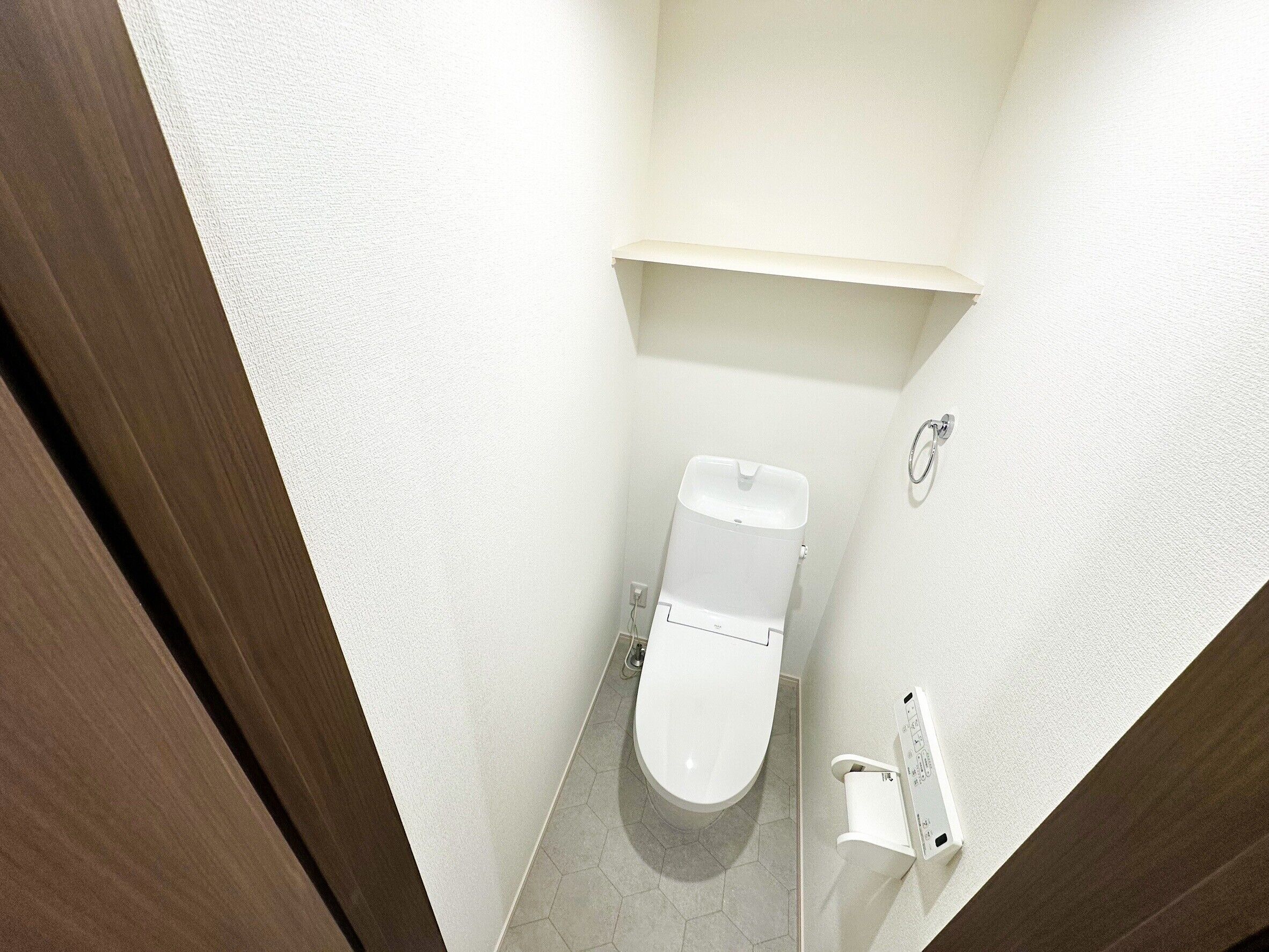 Pieni, kapea kylpyhuone, jossa on valkoinen wc-istuin, wc-paperiteline ja wc-istuimen yläpuolella oleva valkoinen hylly.