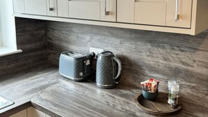 A kitchen with light-colored cabinets, a wooden countertop, a toaster, a kettle, a mug, and a small tray with items. - 0/2 Inchcruin Apartment  - Macaulay Letting (Bonhill)