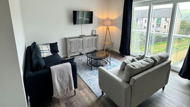 A modern living room with a sofa, a coffee table, and a TV mounted on the wall. There is a window with a view of buildings outside. - Inchconnachan Apartment (Bonhill)