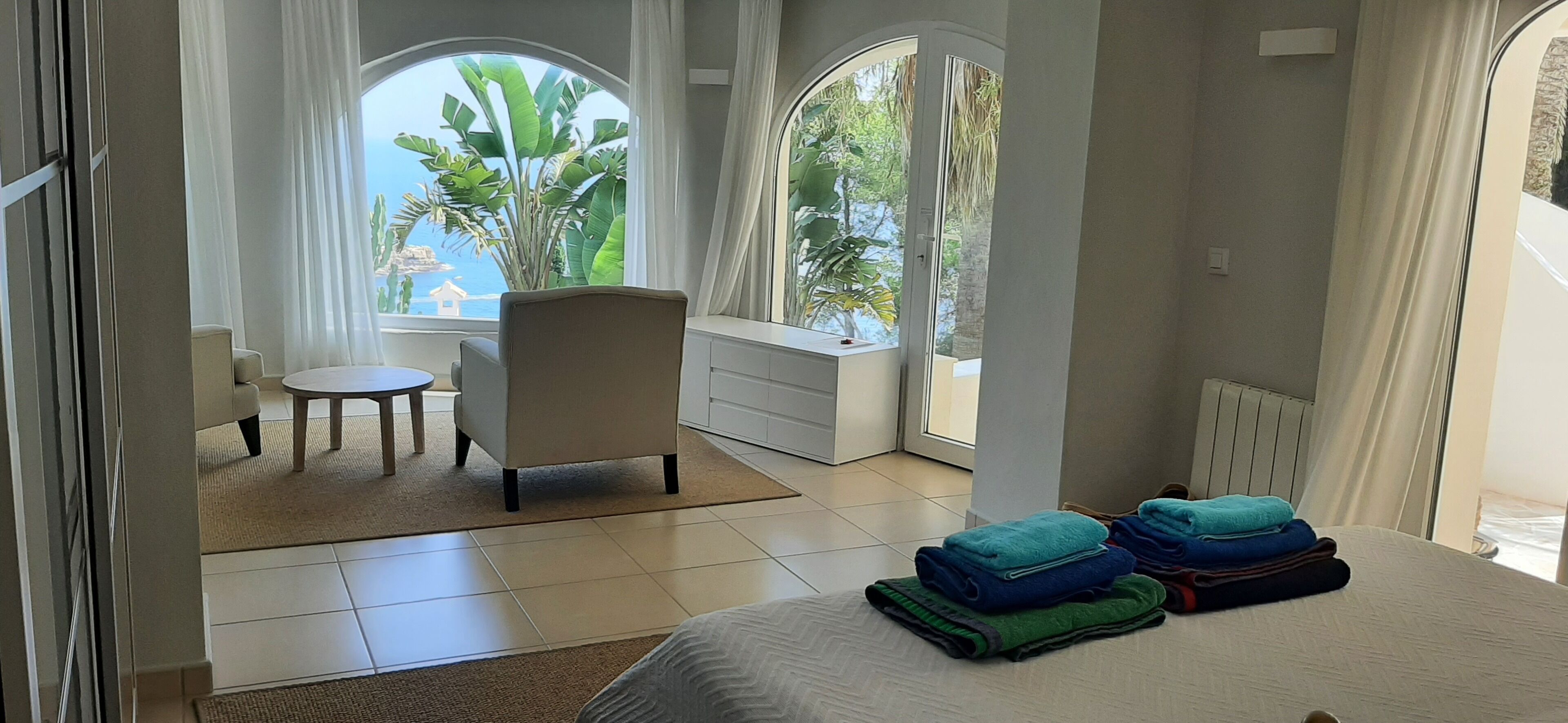 Quarto de hotel com uma cama, uma cadeira, uma mesinha e vista para o oceano através de amplas janelas.