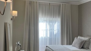 Hotellihuone, jossa on sänky, yöpöytä lampun kanssa, peili ja näkymä ulos ohuiden verhojen läpi.
