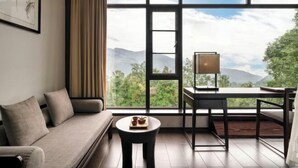 A room with a sofa, a small round table with a tray, a desk with a lamp, and a large window offering a view of greenery and mountains. - Emei Mountain Wannian Xueya Fang Hotel (Emeishan City)