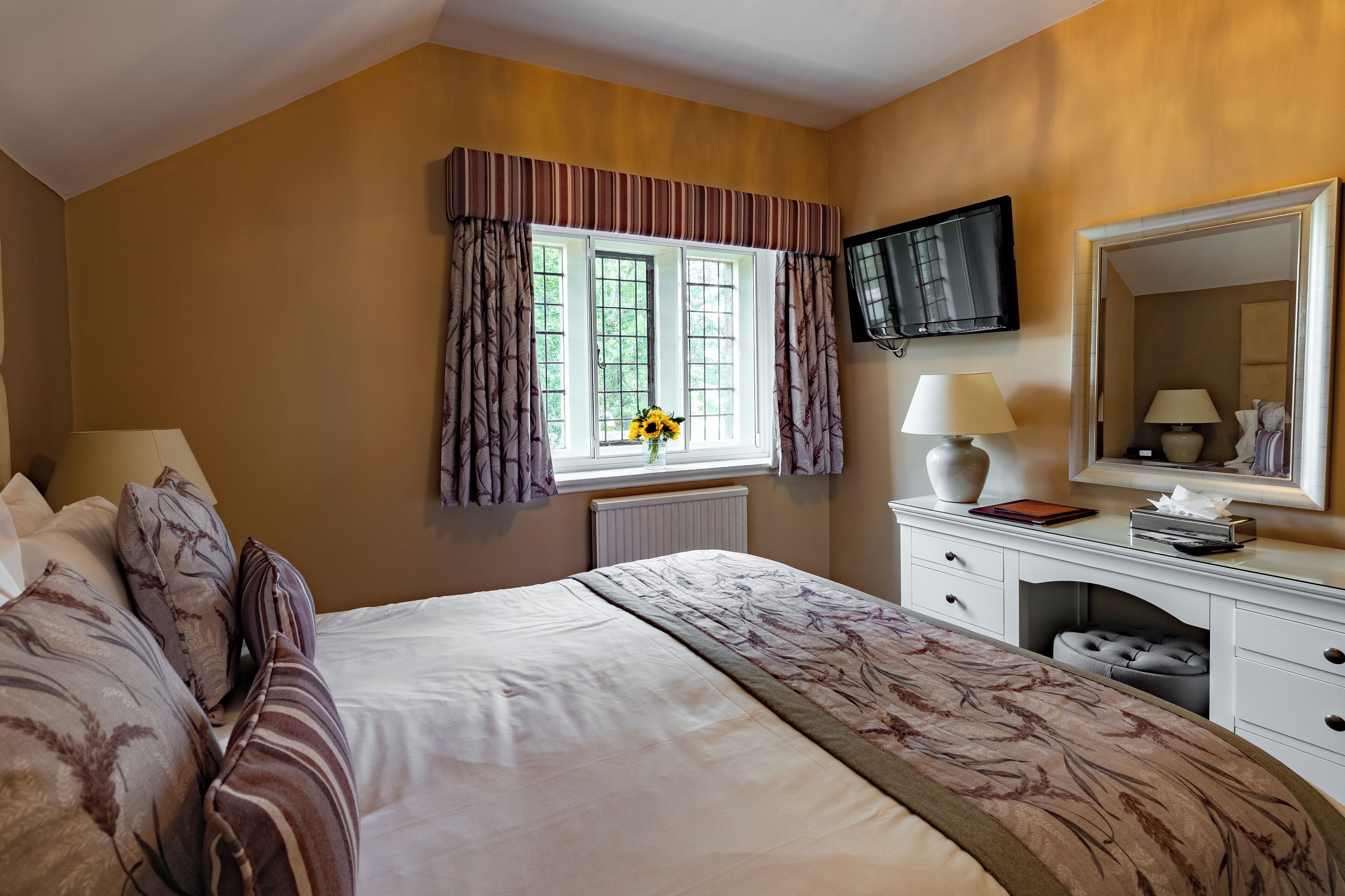 Une chambre d’hôtel avec un grand lit, une commode avec un miroir, une fenêtre avec des rideaux et un téléviseur fixé au mur.