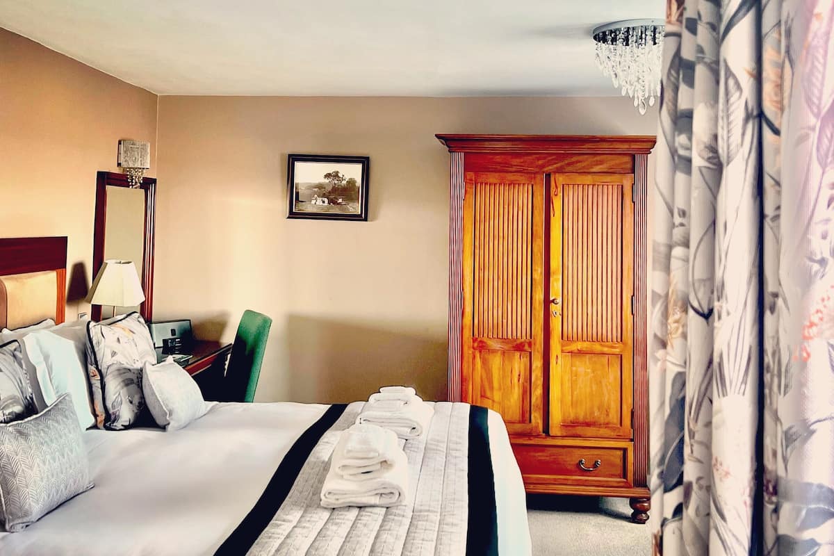 A hotel room with a large bed, a wooden wardrobe, a desk with a lamp, and a framed picture on the wall.