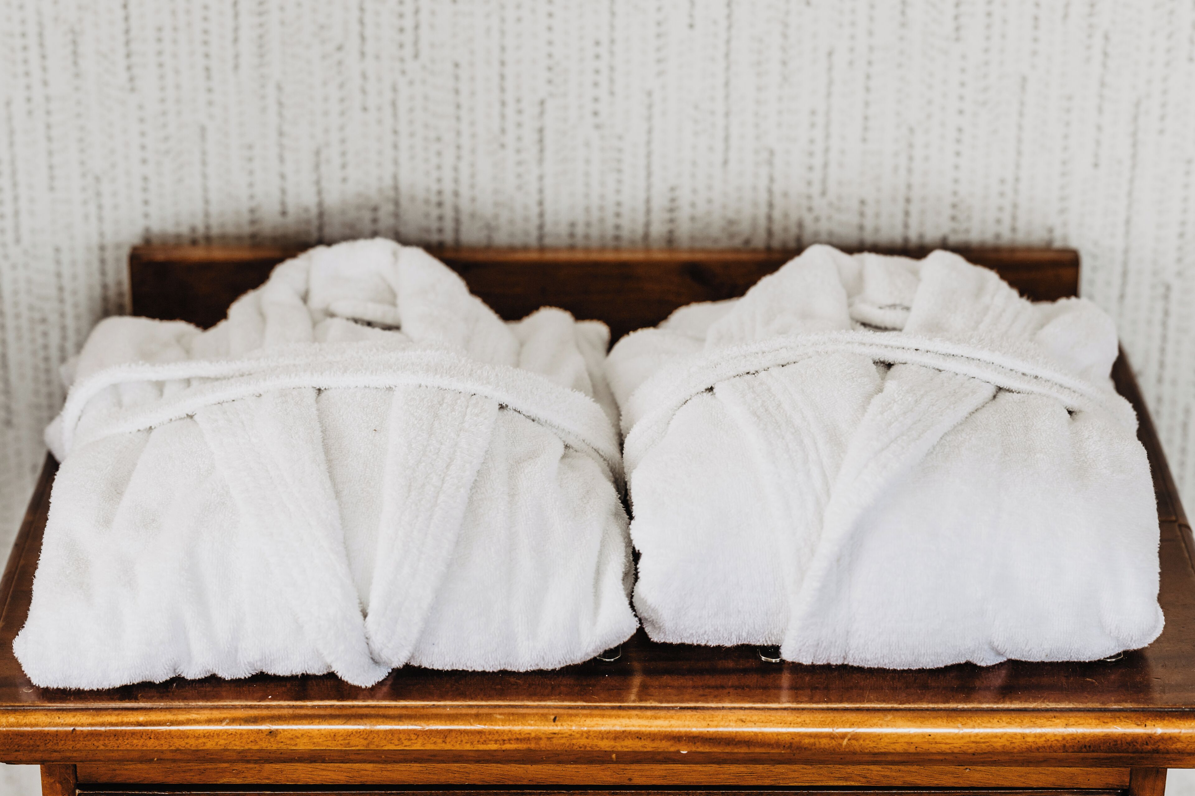 Two white bathrobes on a wooden table against a textured wall.