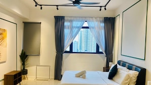 A hotel room with a bed, a ceiling fan, a window with curtains, a radiator, a small plant, and a wall-mounted TV.
