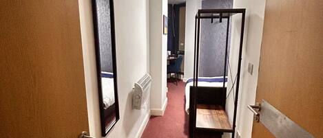 A narrow hallway with a red carpet leading to a room with a bed and a small table.