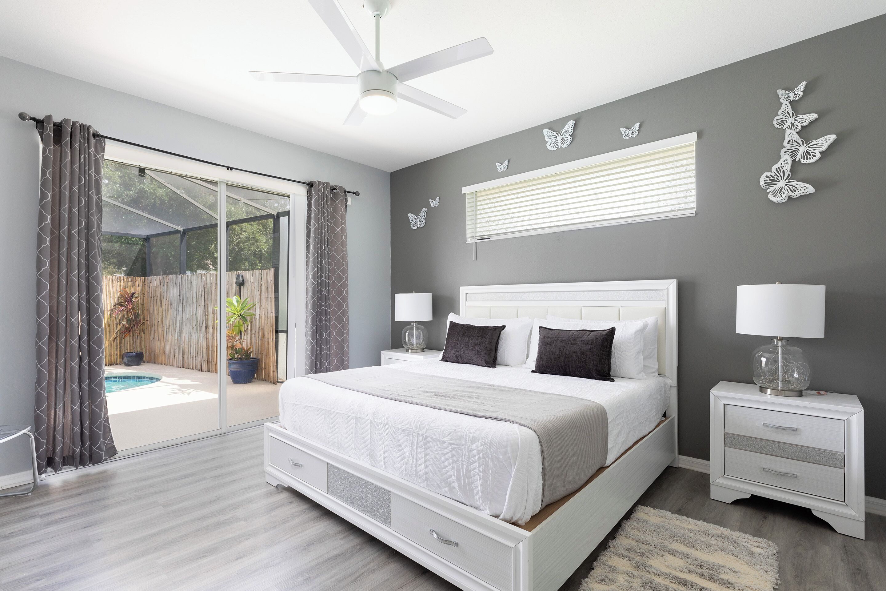 A bedroom with a large bed, a ceiling fan, a nightstand with a lamp, and a view of an outdoor pool area.