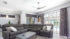 Una sala de estar moderna con un sofá modular, un ventilador de techo y ventanas amplias con cortinas.