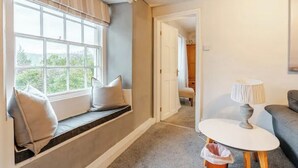 A room with a window seat, a small table with a lamp, and a view of a hallway and another room.