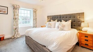 A bedroom with a large bed, a headboard, a bedside table with a lamp, and a window with curtains.
