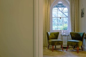 Une pièce avec deux fauteuils verts, une petite table et une grande fenêtre en arc de cercle agrémentée de rideaux.
