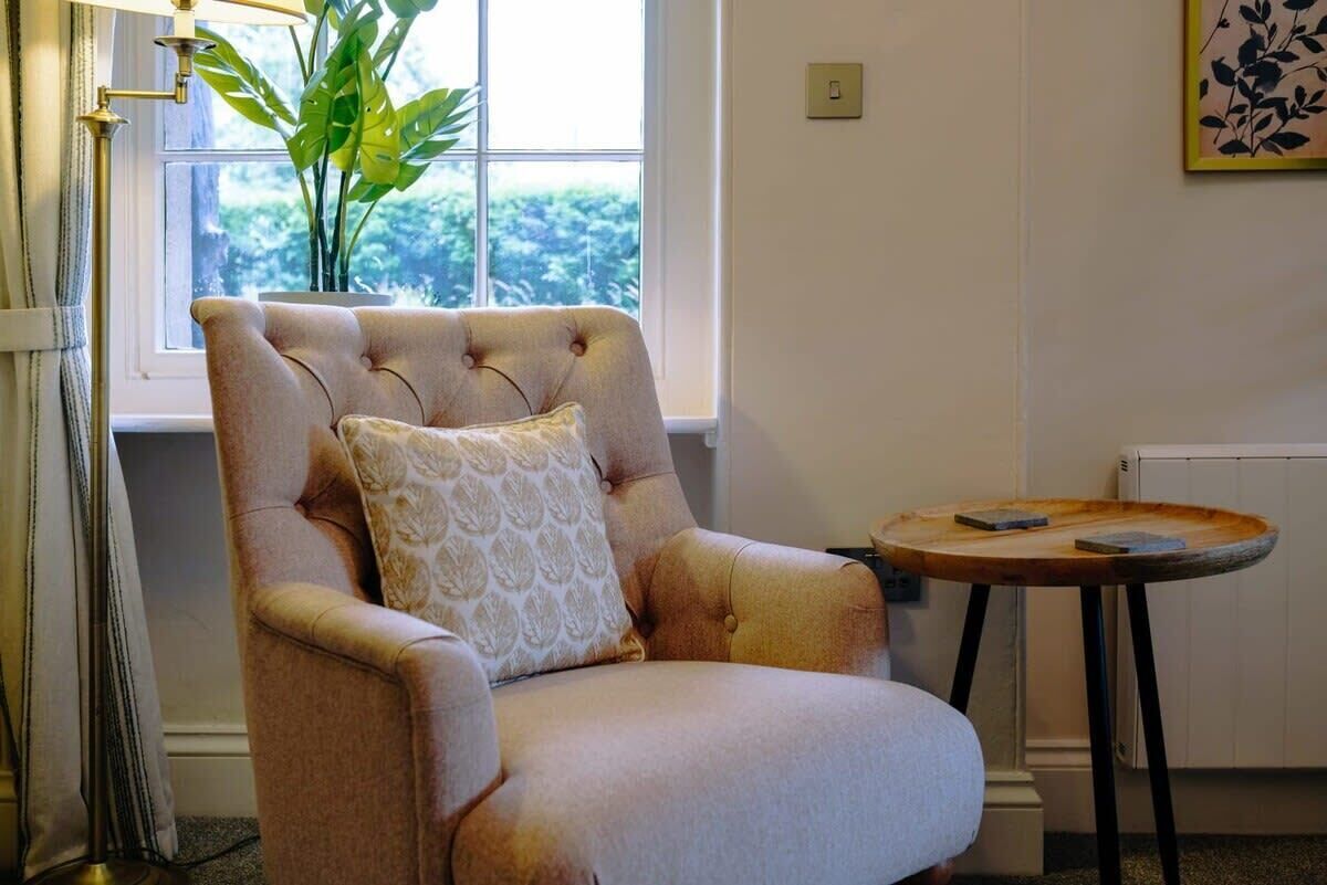 A plush armchair with a patterned cushion, positioned near a window with a view of greenery, a small round table with a book, and a floor lamp.