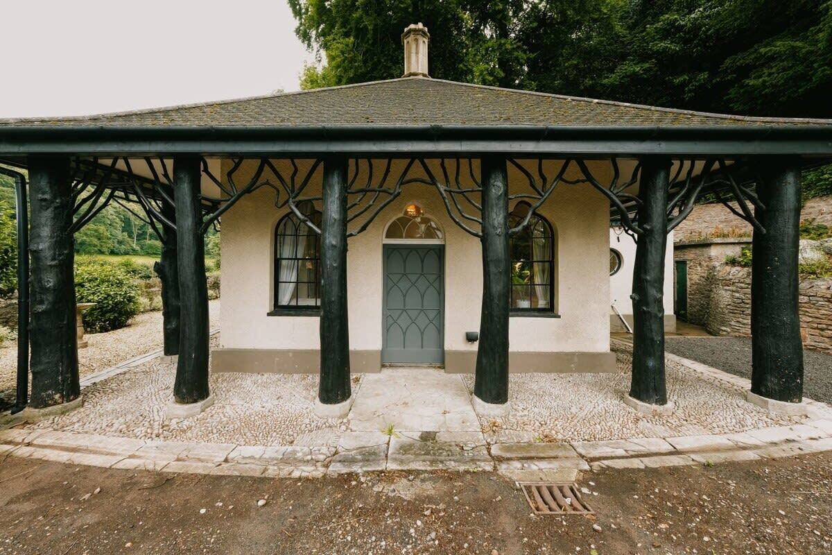 Un edificio pequeño de una sola planta con techo inclinado y una puerta central, rodeado por un camino de grava y sostenido por columnas negras.