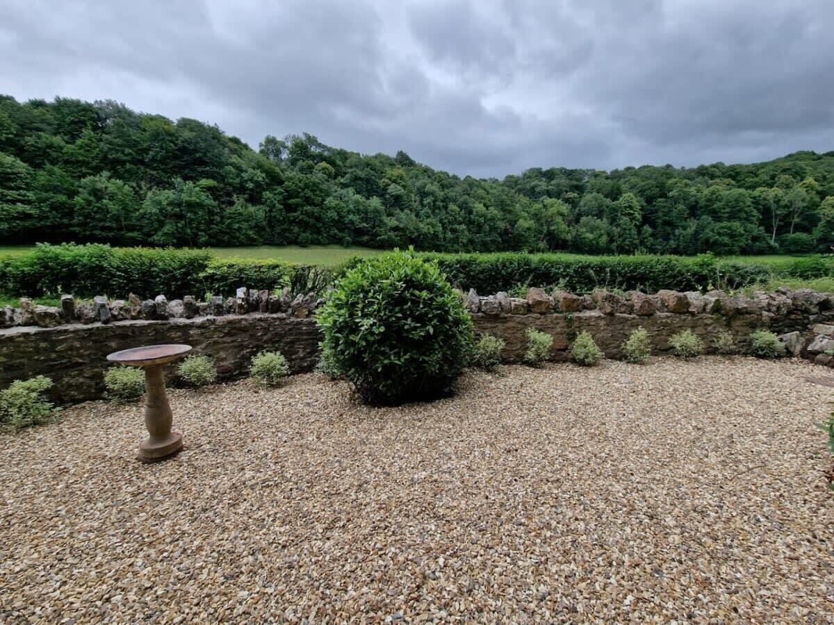 Ein Garten mit einem Steintisch und einem Busch, umgeben von einer Steinmauer und einem Kiesweg.