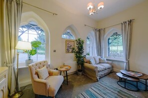 A cozy living room with a sofa, armchair, and coffee table, large windows with curtains, and a chandelier. - The Lodge Gatehouse at Coombe Lodge (Bristol)