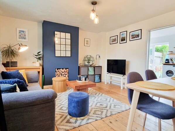 A modern living room with a sofa, ottoman, coffee table, and TV. There is a dining area with chairs and a table. The room has a blue accent wall and a large window.