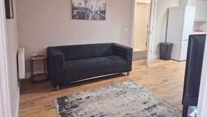 Una sala de estar con un sofá, una mesita auxiliar, una alfombra, un cuadro de un paisaje urbano y una puerta que da a una cocina.
