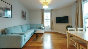A modern living room with a sofa, a small table, a dining table with chairs, a flat-screen TV mounted on the wall, and a large window with curtains.