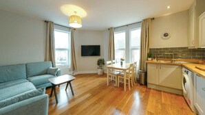 A modern apartment with a living room, dining area, and kitchen. It features hardwood flooring, a flat-screen TV, a dining table with chairs, a sofa, and a kitchen with a washing machine.