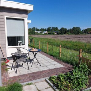 A patio with outdoor furniture, a view of a field, and a house in the background. - Modern Bungalow Close to Beach With Views (Sint Maartensvlotbrug)
