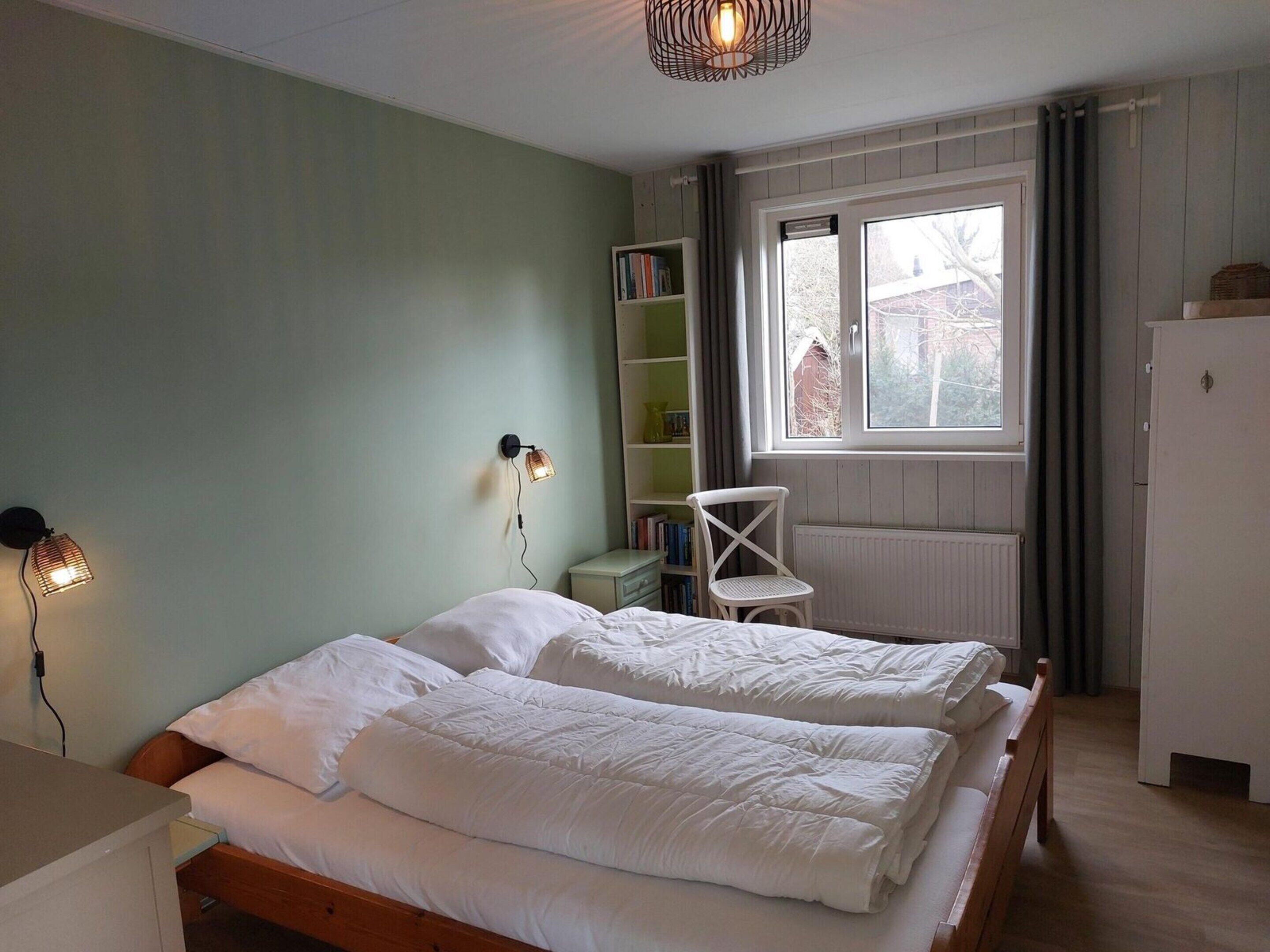 A bedroom with two beds, a bookshelf, a chair, and a window with a view of buildings outside.