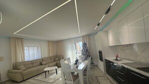 A modern living room with a beige sofa, a dining table with chairs, a kitchen area with dark cabinets, and a Christmas tree.