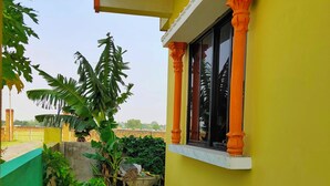 A yellow building with a window and orange columns, a concrete pathway, and green plants. - Tathagata Homestay (Lumbini)