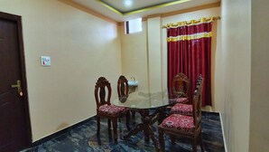 A conference room with a round table, four wooden chairs with red and white patterned cushions, a ceiling fan, recessed lighting, a red curtain with gold trim, and a marble floor. - Tathagata Homestay (Lumbini)