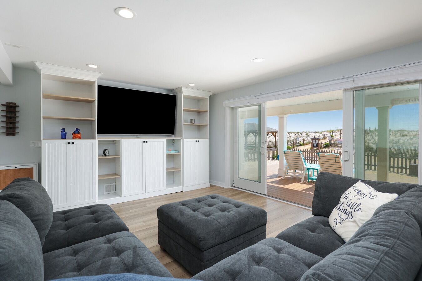 A modern living room with a flat-screen TV, grey sofas, and a sliding glass door leading to a balcony with a view of the beach.
